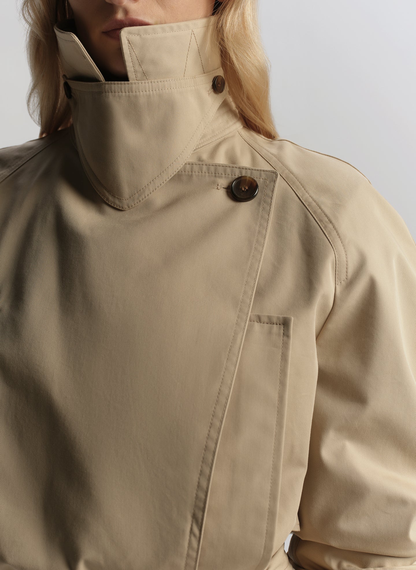 Woman wearing a beige trench coat, white shirt, and blue jeans on a plain background