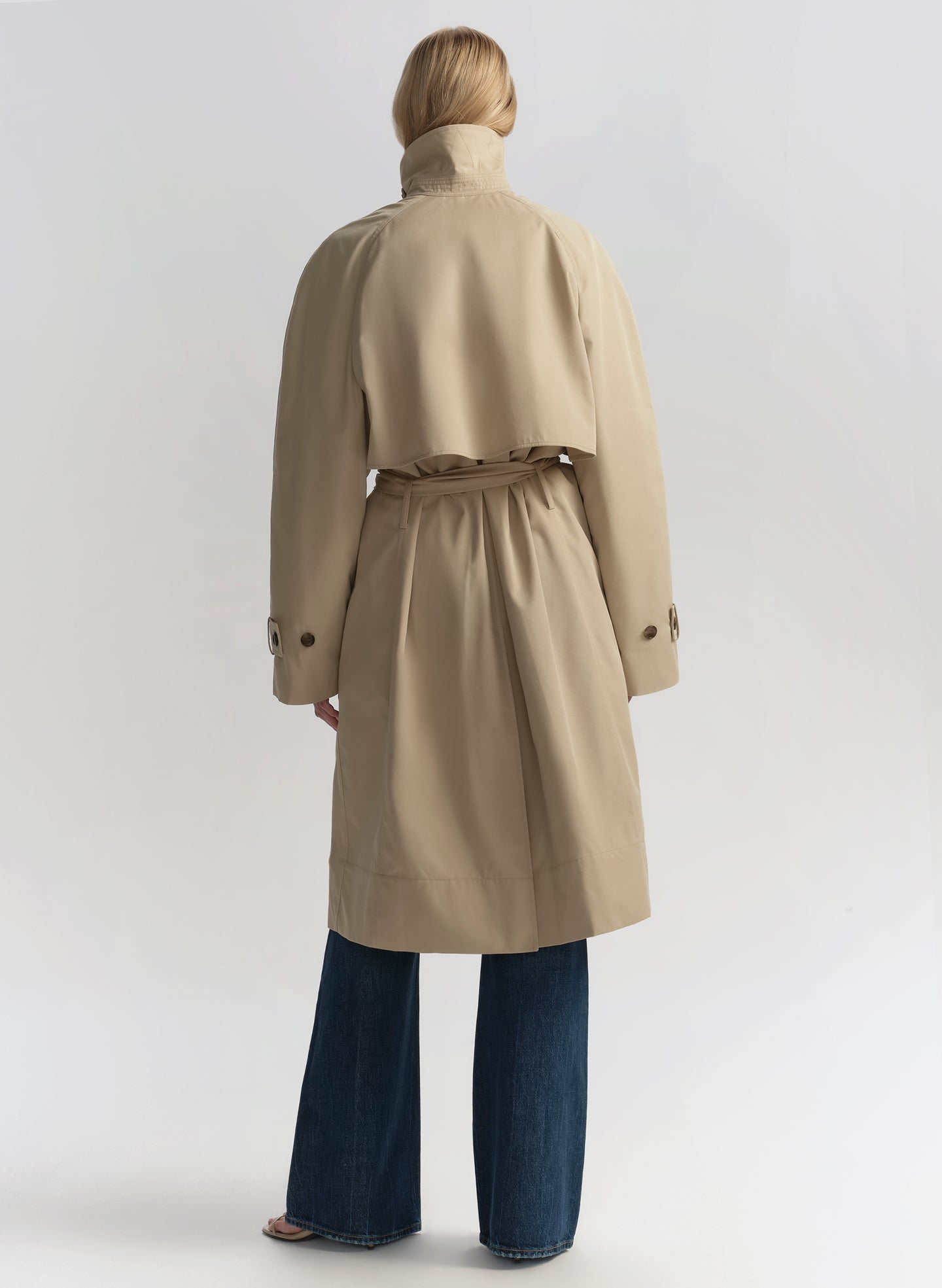 Woman wearing a beige trench coat, white shirt, and blue jeans on a plain background