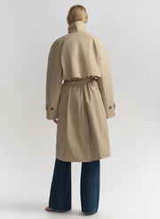 Woman wearing a beige trench coat, white shirt, and blue jeans on a plain background