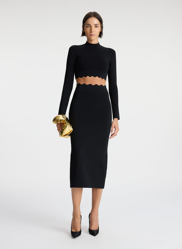 front view of woman wearing scalloped trim long sleeve black crop top and matching black skirt