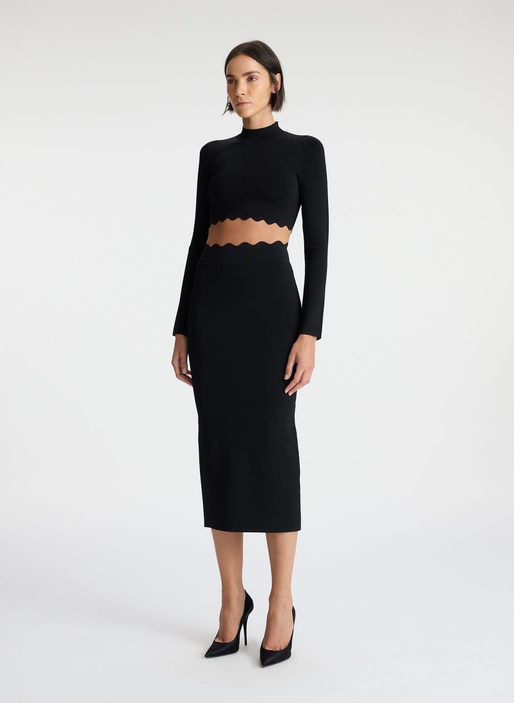 side view of woman wearing black long sleeve crop top with scallop detailing and matching black scalloped detailing knit midi skirt