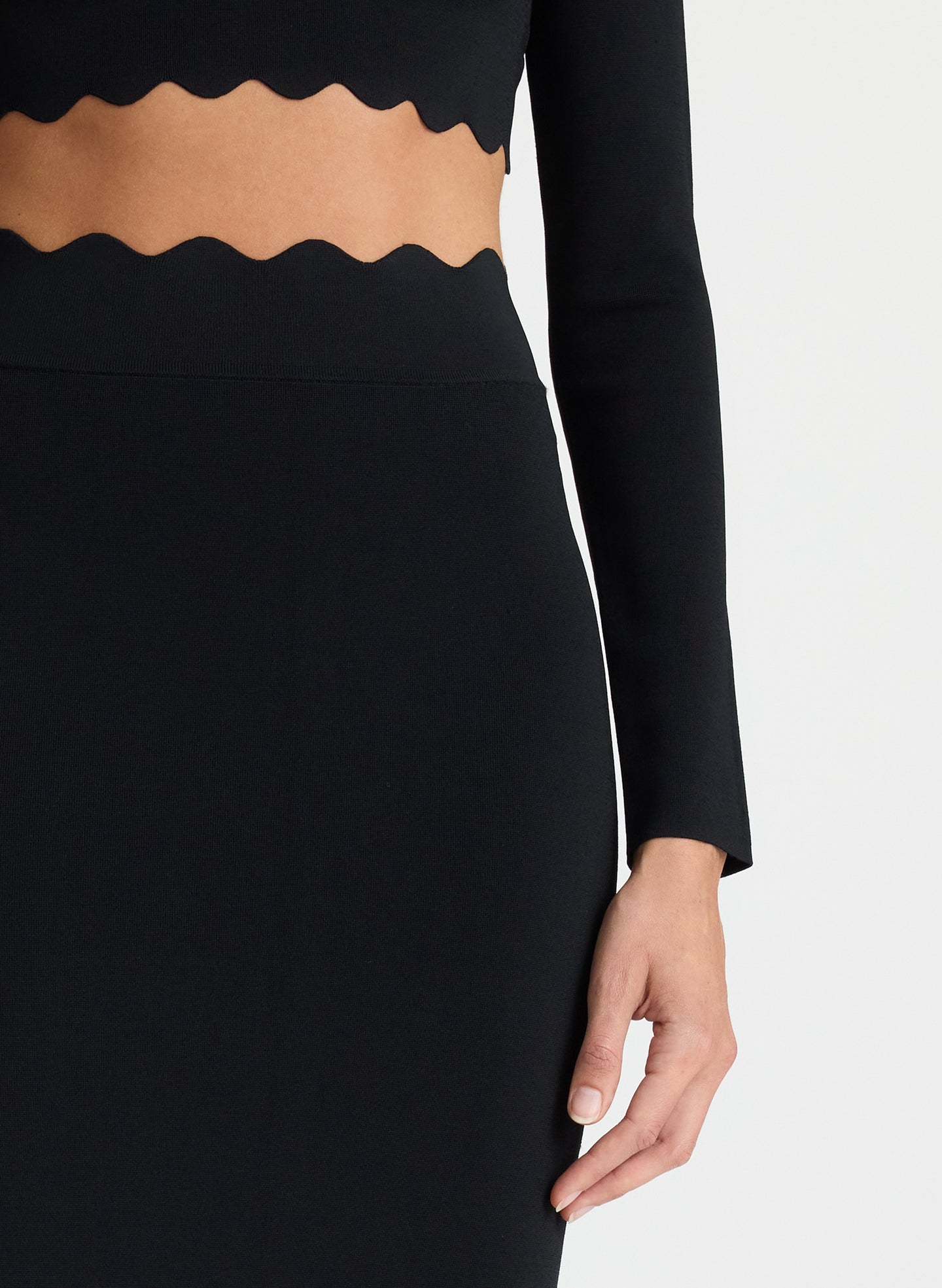 detail view of woman wearing black long sleeve crop top with scallop detailing and matching black scalloped detailing knit midi skirt