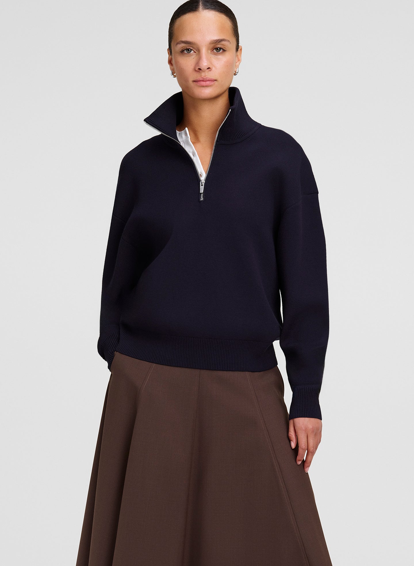 A woman modeling a dark navy blue quarter-zip sweater with a stand-up collar, paired with a brown skirt, against a light gray studio background.
