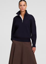 A woman modeling a dark navy blue quarter-zip sweater with a stand-up collar, paired with a brown skirt, against a light gray studio background.