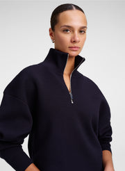 A model wearing a dark navy blue quarter-zip knit sweater with a high ribbed mock neck collar, photographed against a neutral light gray studio background.