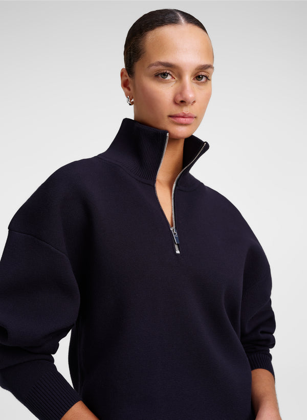A model wearing a dark navy blue quarter-zip knit sweater with a high ribbed mock neck collar, photographed against a neutral light gray studio background.