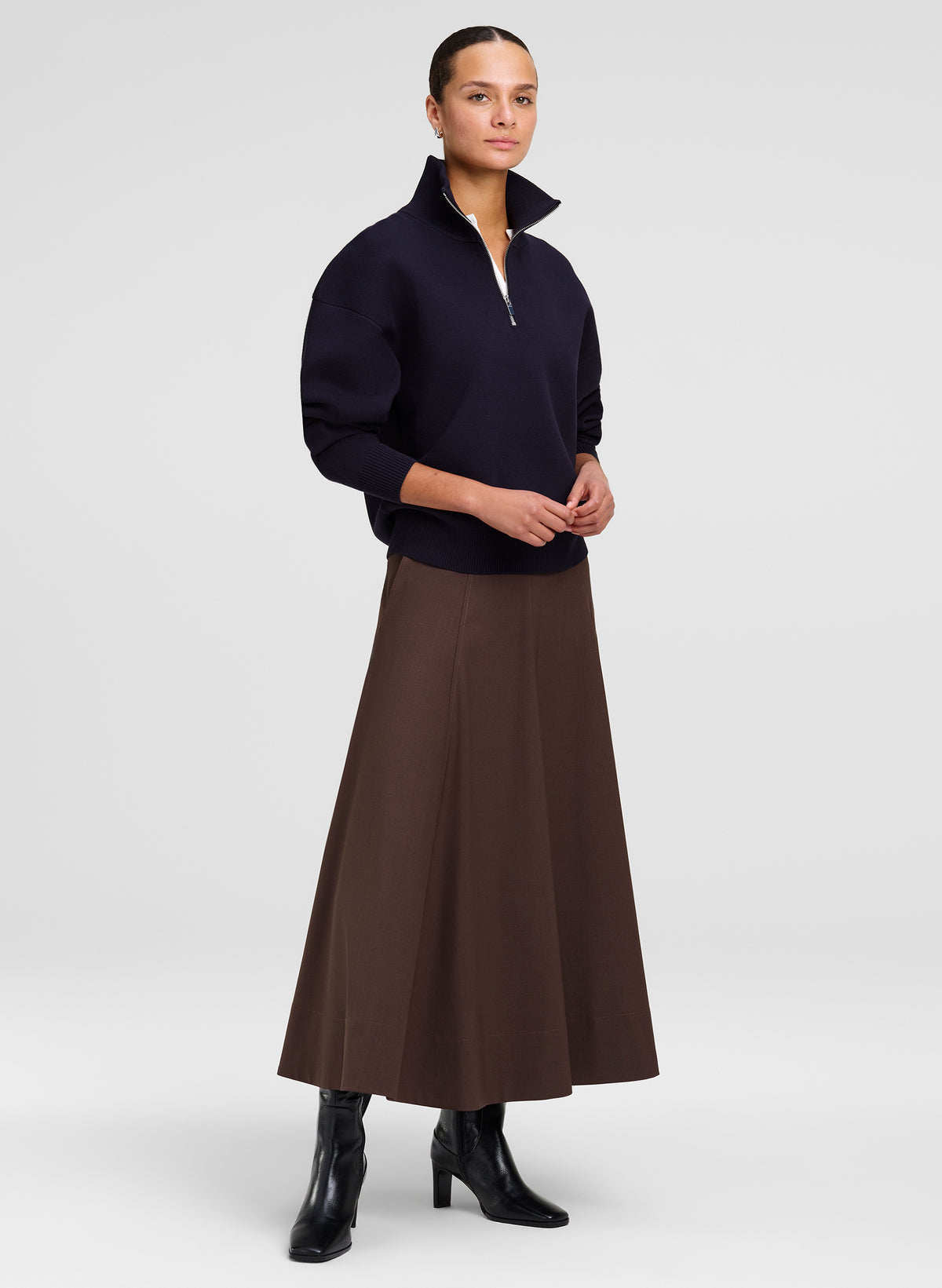 A model stands against a light gray studio background wearing a dark navy quarter-zip mock-neck sweater, a brown A-line midi skirt, and black ankle boots.