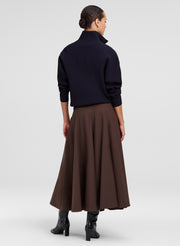 A model shown from the back wearing a dark navy mock-neck knit sweater and a brown pleated midi skirt, paired with black heeled boots, against a light gray studio background.