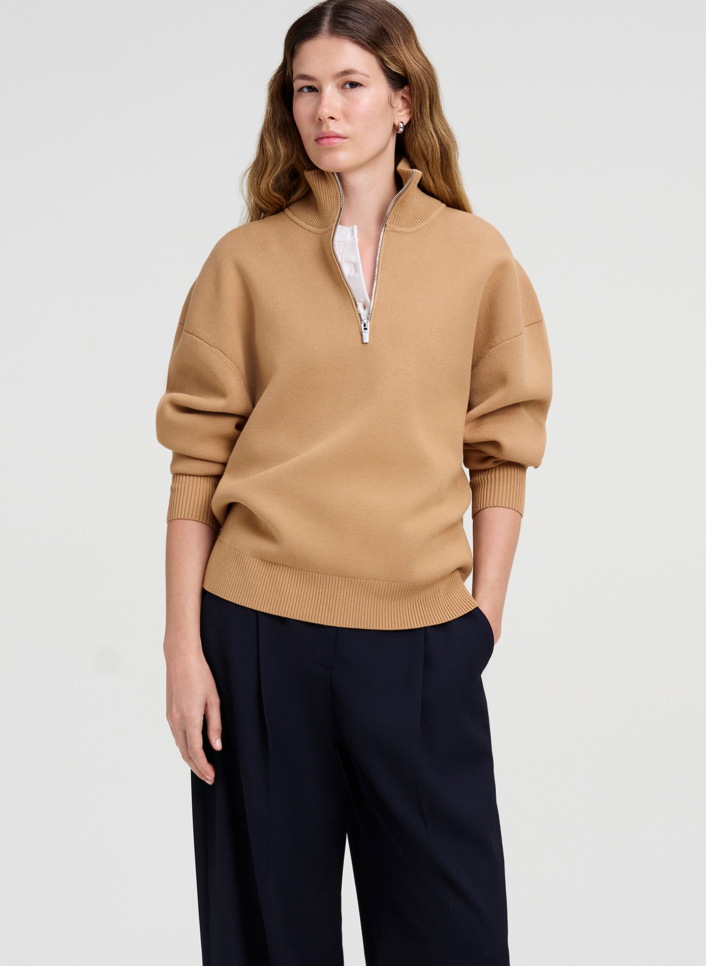 A woman modeling a camel-colored quarter-zip knit sweater with a white inner collar detail, paired with dark navy wide-leg trousers, against a light gray studio background.