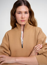 A woman wearing a camel-colored ribbed knit half-zip sweater with a stand-up collar, photographed against a neutral light gray studio background.