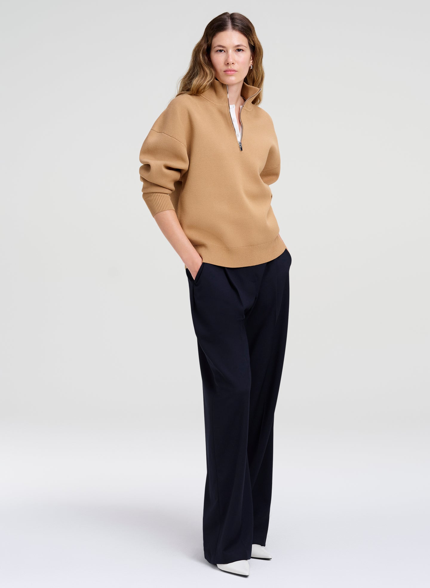 A woman modeling a camel-colored half-zip sweater with voluminous sleeves and dark navy wide-leg trousers, standing against a light gray studio background.