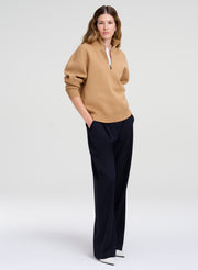 A woman modeling a camel-colored half-zip sweater with voluminous sleeves and dark navy wide-leg trousers, standing against a light gray studio background.