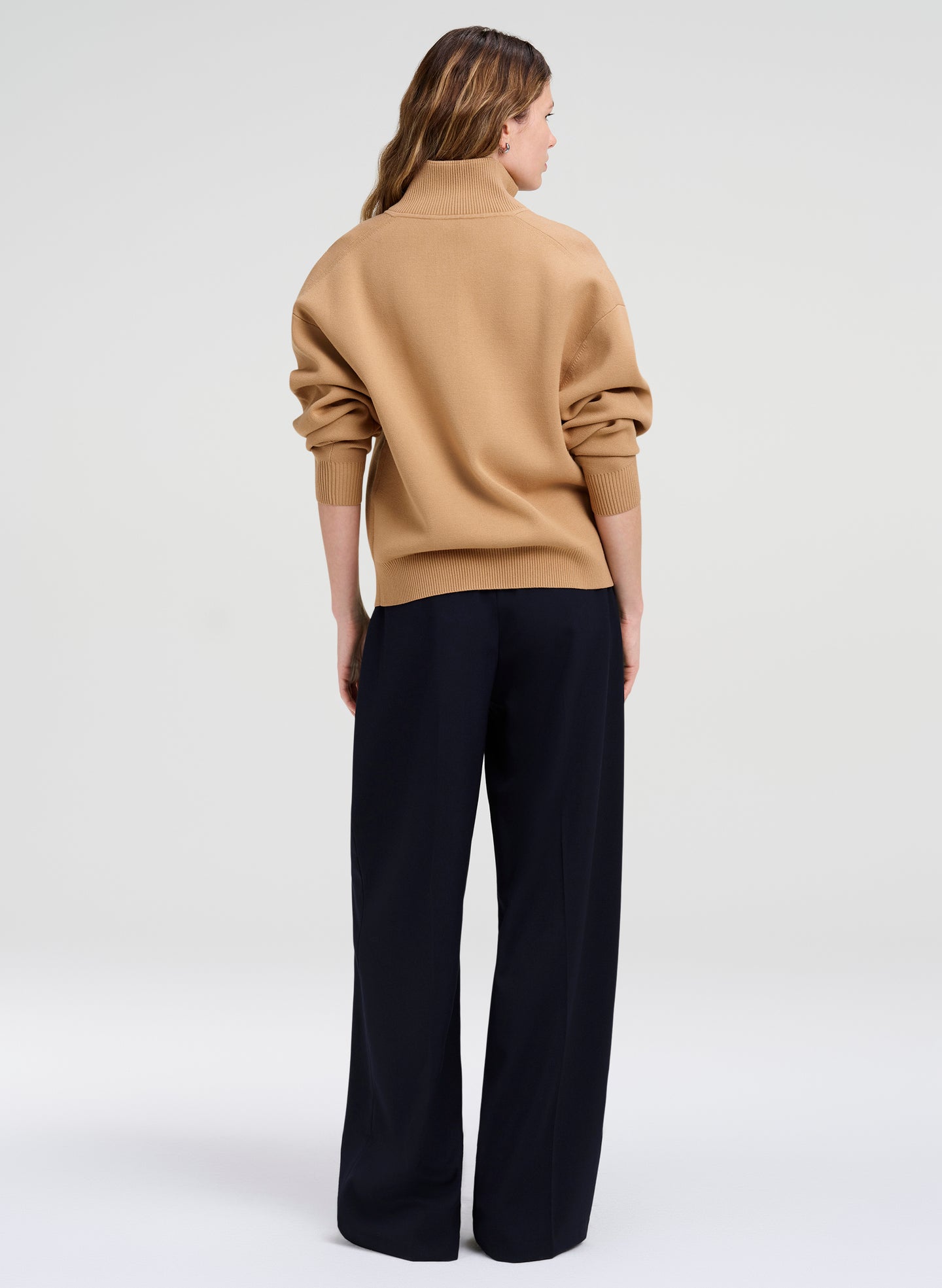 A woman shown from the back wearing a camel-colored knit mock-neck sweater and dark navy wide-leg trousers, photographed against a seamless light gray studio background.
