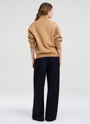 A woman shown from the back wearing a camel-colored knit mock-neck sweater and dark navy wide-leg trousers, photographed against a seamless light gray studio background.