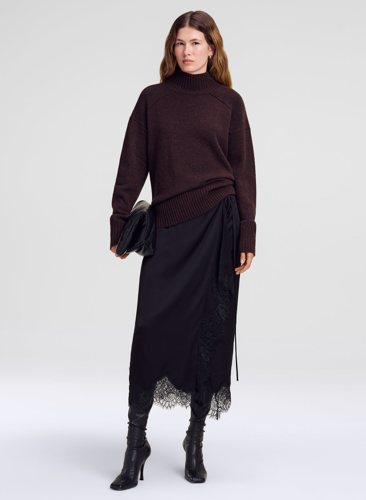 A model stands against a light gray studio background wearing a dark brown ribbed turtleneck sweater, a black midi skirt with a lace hem, and black heeled ankle boots, while holding a black clutch.