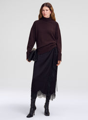 A model stands against a light gray studio background wearing a dark brown ribbed turtleneck sweater, a black midi skirt with a lace hem, and black heeled ankle boots, while holding a black clutch.