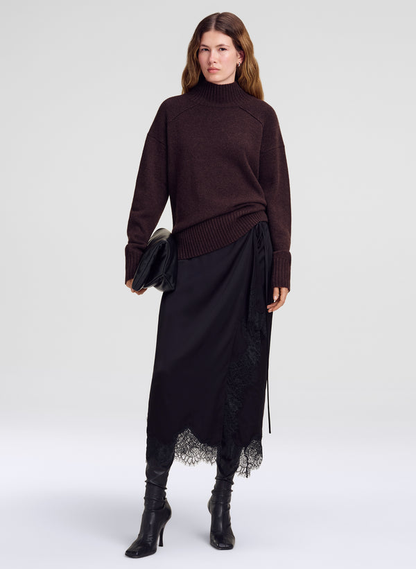 A model stands against a light gray studio background wearing a dark brown ribbed turtleneck sweater, a black midi skirt with a lace hem, and black heeled ankle boots, while holding a black clutch.