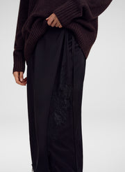 A close-up of a person wearing  a black skirt with a sheer black lace panel on the side, paired with a dark brown ribbed knit sweater, against a light gray studio background.