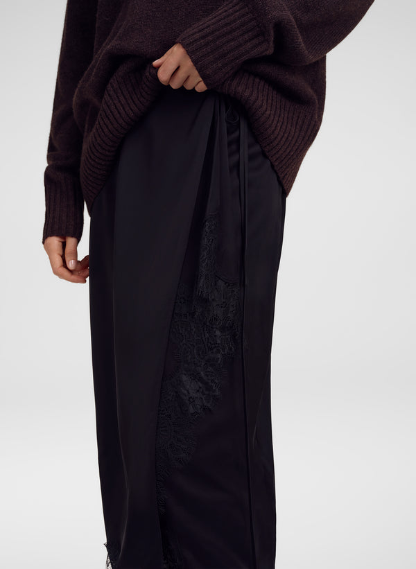A close-up of a person wearing  a black skirt with a sheer black lace panel on the side, paired with a dark brown ribbed knit sweater, against a light gray studio background.