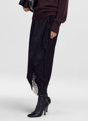 A model wearing a black draped wrap skirt with lace trim, paired with a dark brown ribbed sweater and black pointed-toe heeled boots, against a light gray studio background.