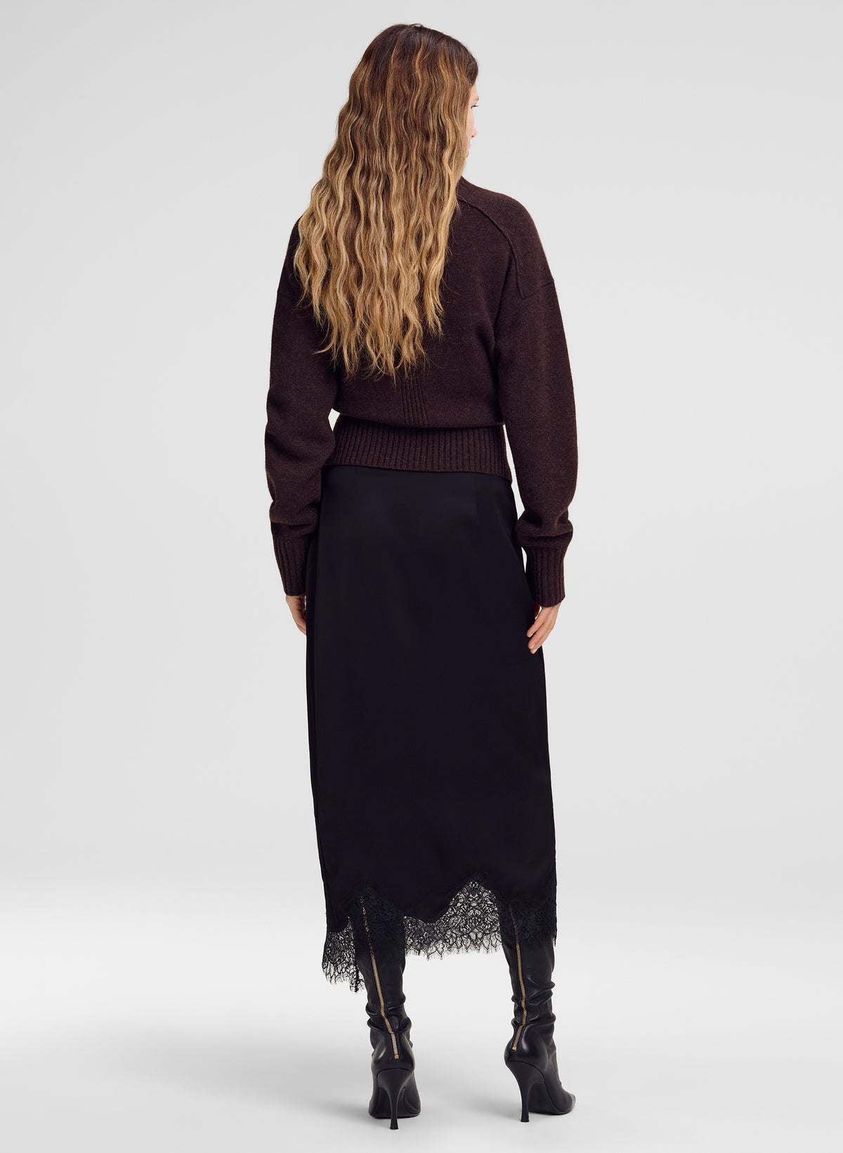 A woman shown from the back wearing a dark brown knit sweater and a long black skirt with a lace hem, paired with black heeled boots, against a light gray studio background.
