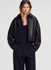 A woman modeling a cropped black leather jacket with a collar and front buttons, worn over a dark top and dark trousers, against a light gray studio background.