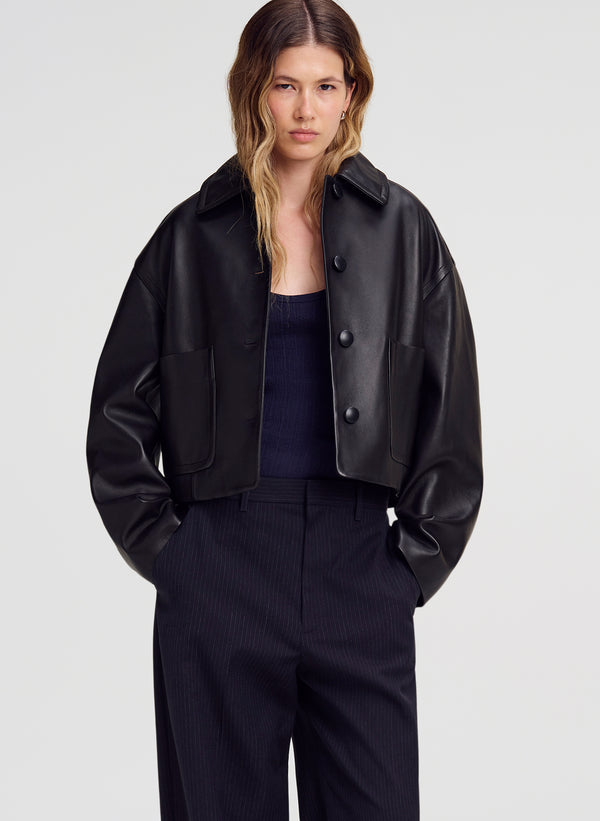 A woman modeling a cropped black leather jacket with a collar and front buttons, worn over a dark top and dark trousers, against a light gray studio background.