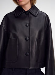 A close-up of a model wearing a black leather jacket with a wide collar, three large front buttons, and two large front patch pockets.