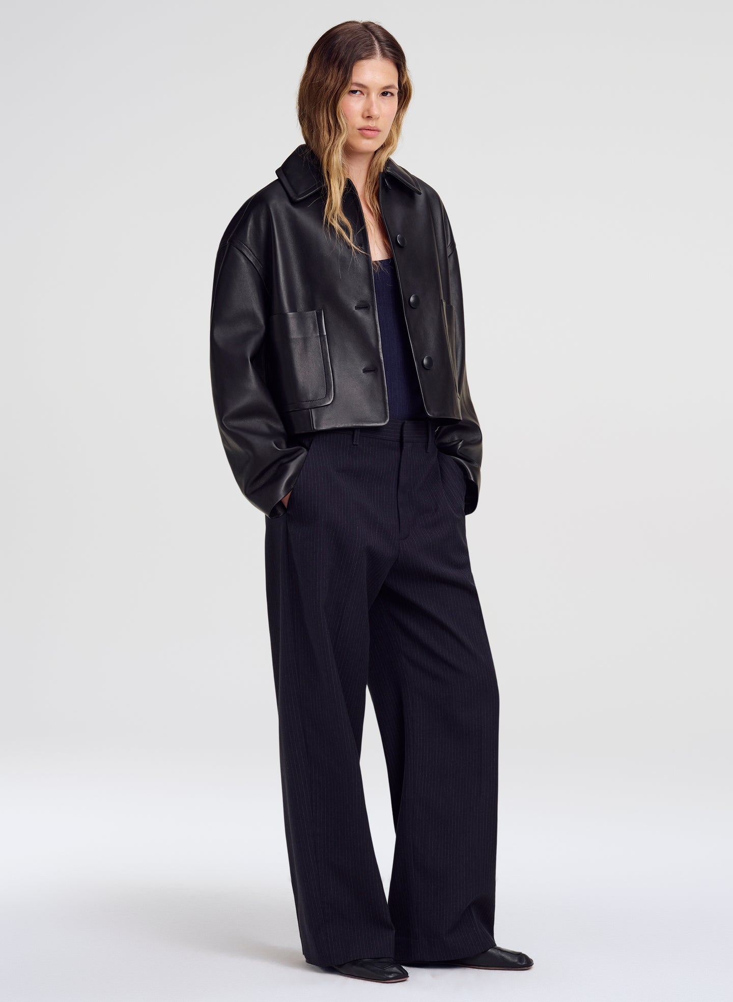 A model stands against a light gray studio background wearing a cropped black leather jacket over a dark top and black wide-leg trousers.