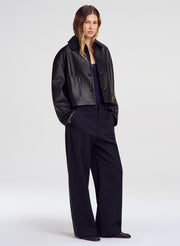 A model stands against a light gray studio background wearing a cropped black leather jacket over a dark top and black wide-leg trousers.