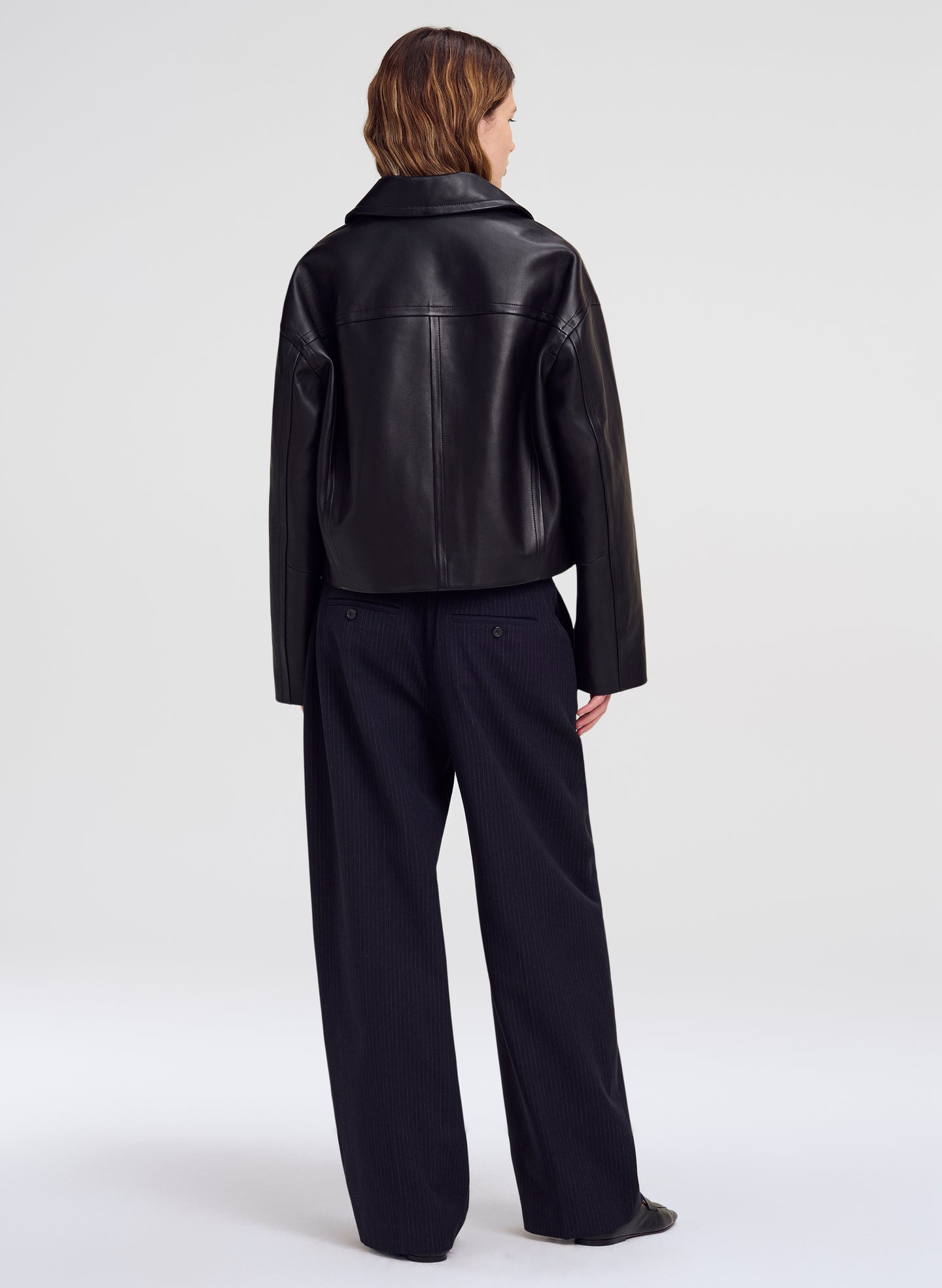 A model shown from the back wearing a black leather jacket and dark wide-leg trousers against a light gray studio background.
