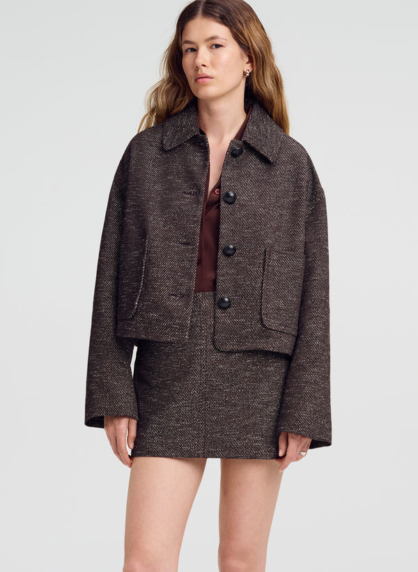 A woman modeling a dark brown tweed cropped jacket and matching mini skirt set against a light gray studio background.
