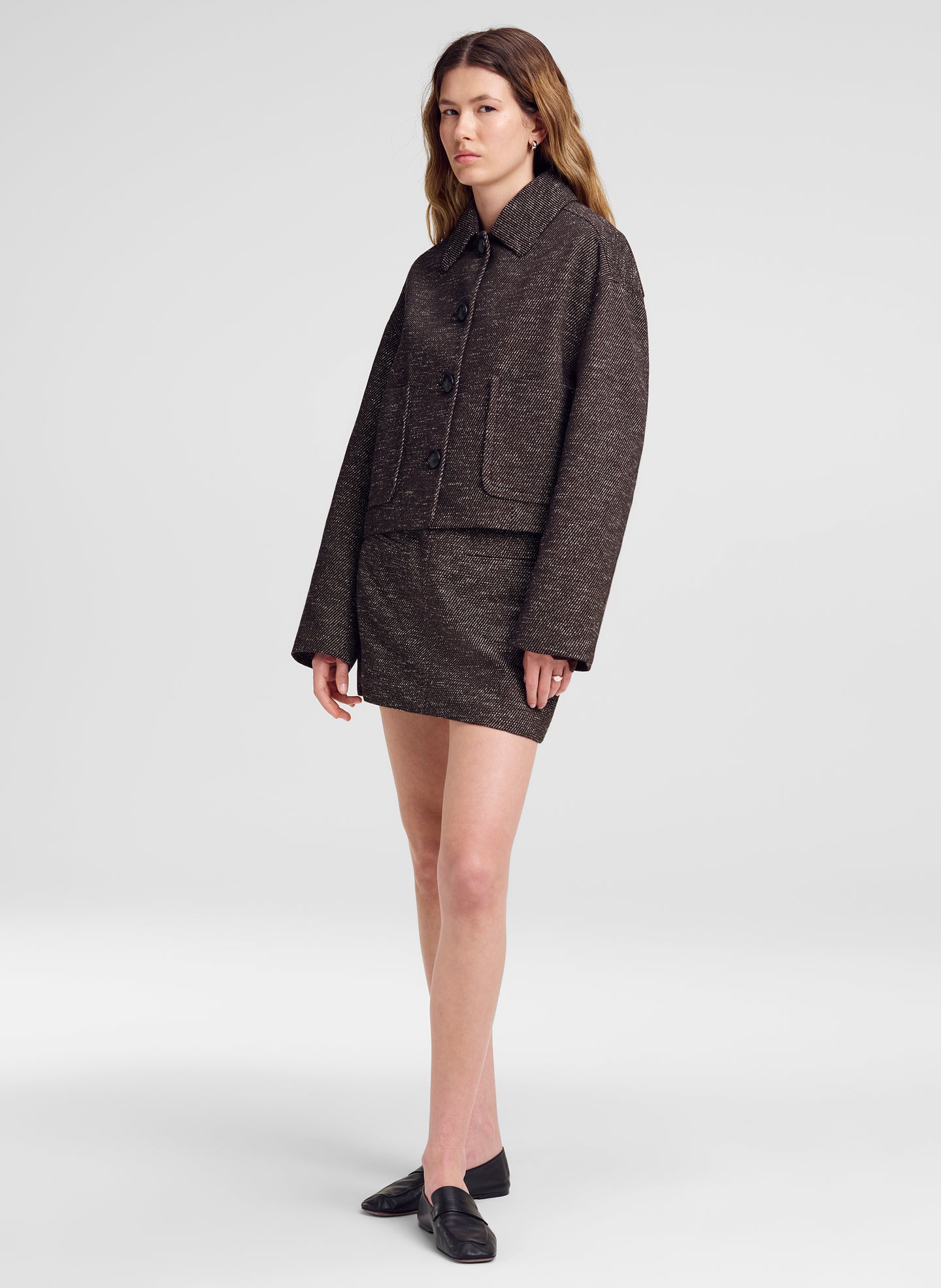 A model stands against a light gray studio background wearing a dark brown tweed jacket and matching mini skirt set, paired with black loafers.