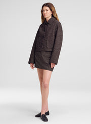A model stands against a light gray studio background wearing a dark brown tweed jacket and matching mini skirt set, paired with black loafers.
