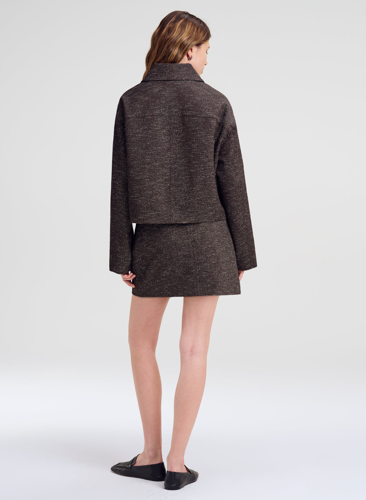 A model shown from the back wearing a dark brown tweed cropped jacket and matching mini skirt, paired with black flat shoes, against a light gray studio background.