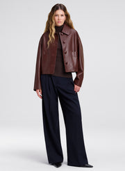 A model stands against a light gray studio background wearing a cropped brown leather jacket over a dark turtleneck, paired with dark wide-leg trousers and black shoes.