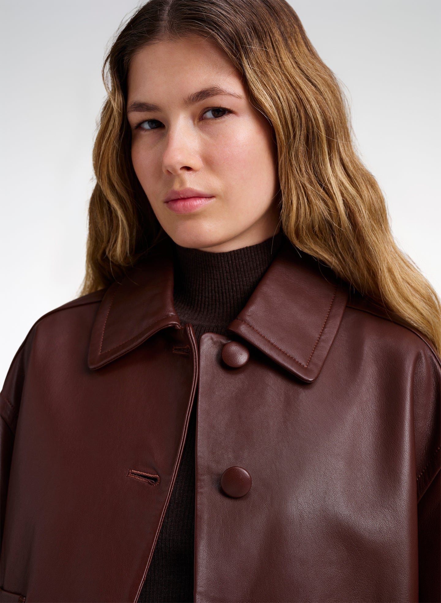 A close-up of a woman wearing a dark brown leather jacket with a wide collar and large buttons over a black turtleneck, against a light gray studio background.