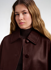 A close-up of a woman wearing a dark brown leather jacket over a black turtleneck sweater, set against a plain light gray studio background.
