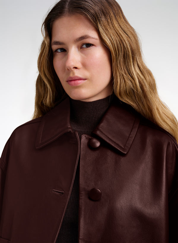 A close-up of a woman wearing a dark brown leather jacket over a black turtleneck sweater, set against a plain light gray studio background.