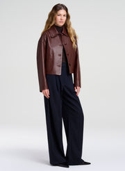 A woman modeling a cropped brown leather jacket with a collar and front buttons, worn over a brown top and paired with dark wide-leg trousers, against a light gray studio background.