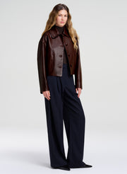 A woman stands against a light gray studio background wearing a cropped dark brown leather jacket over a black top and dark navy wide-leg trousers.