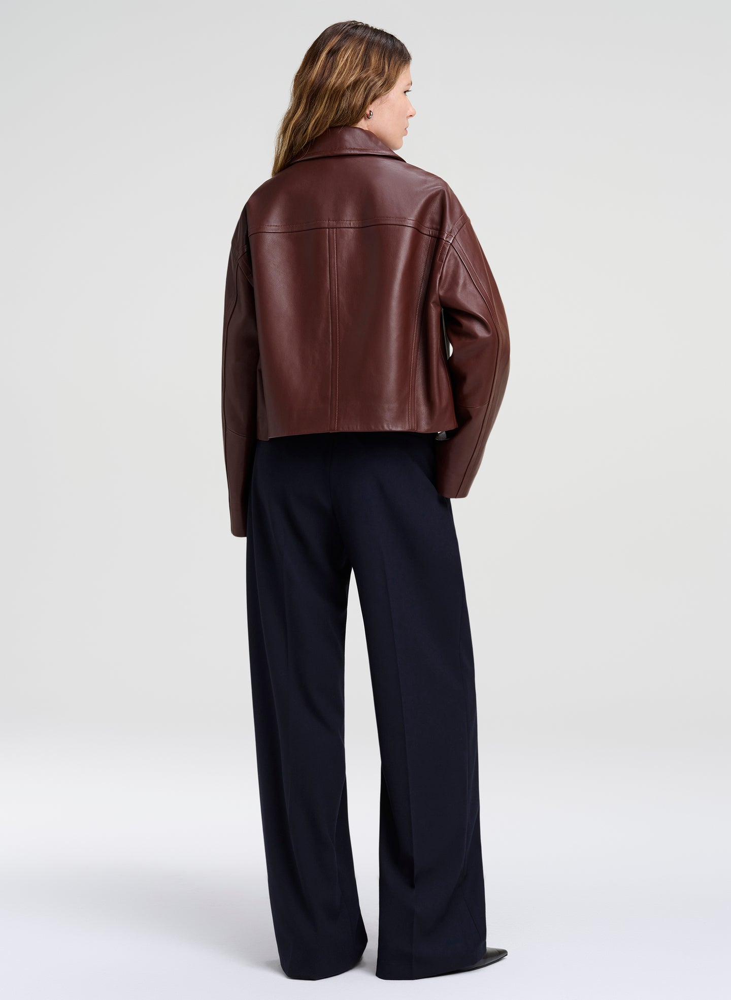 A woman shown from the back wearing a cropped brown leather jacket and dark wide-leg trousers, photographed against a clean light gray studio background.
