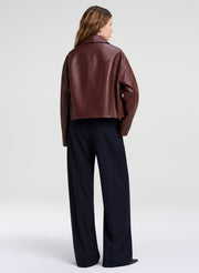 A woman shown from the back wearing a cropped brown leather jacket and dark wide-leg trousers, photographed against a clean light gray studio background.