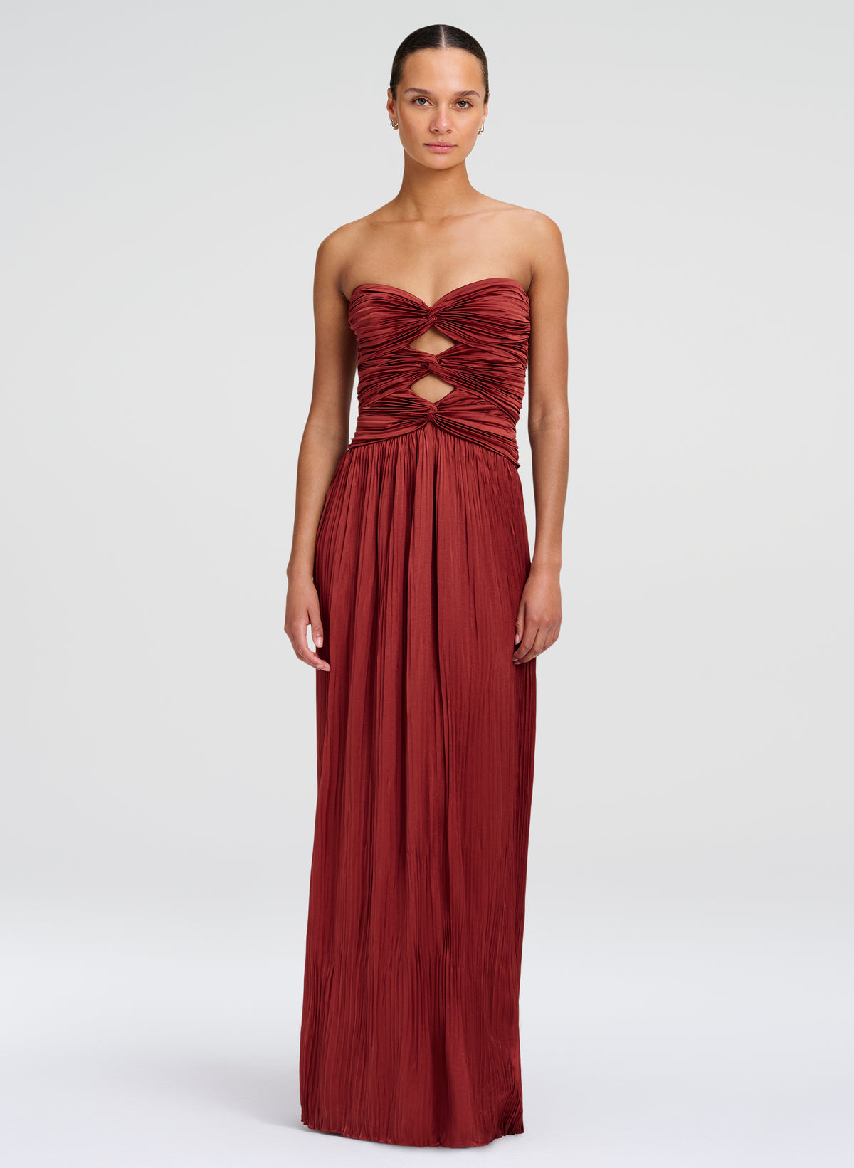A woman modeling a strapless floor-length gown in a deep burgundy red. The dress features a ruched, crisscrossed bodice with cutout details at the waist and a flowing pleated skirt, set against a plain light gray studio background.