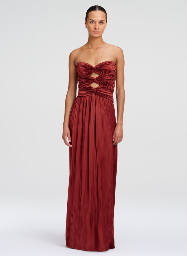 A woman modeling a strapless floor-length gown in a deep burgundy red. The dress features a ruched, crisscrossed bodice with cutout details at the waist and a flowing pleated skirt, set against a plain light gray studio background.