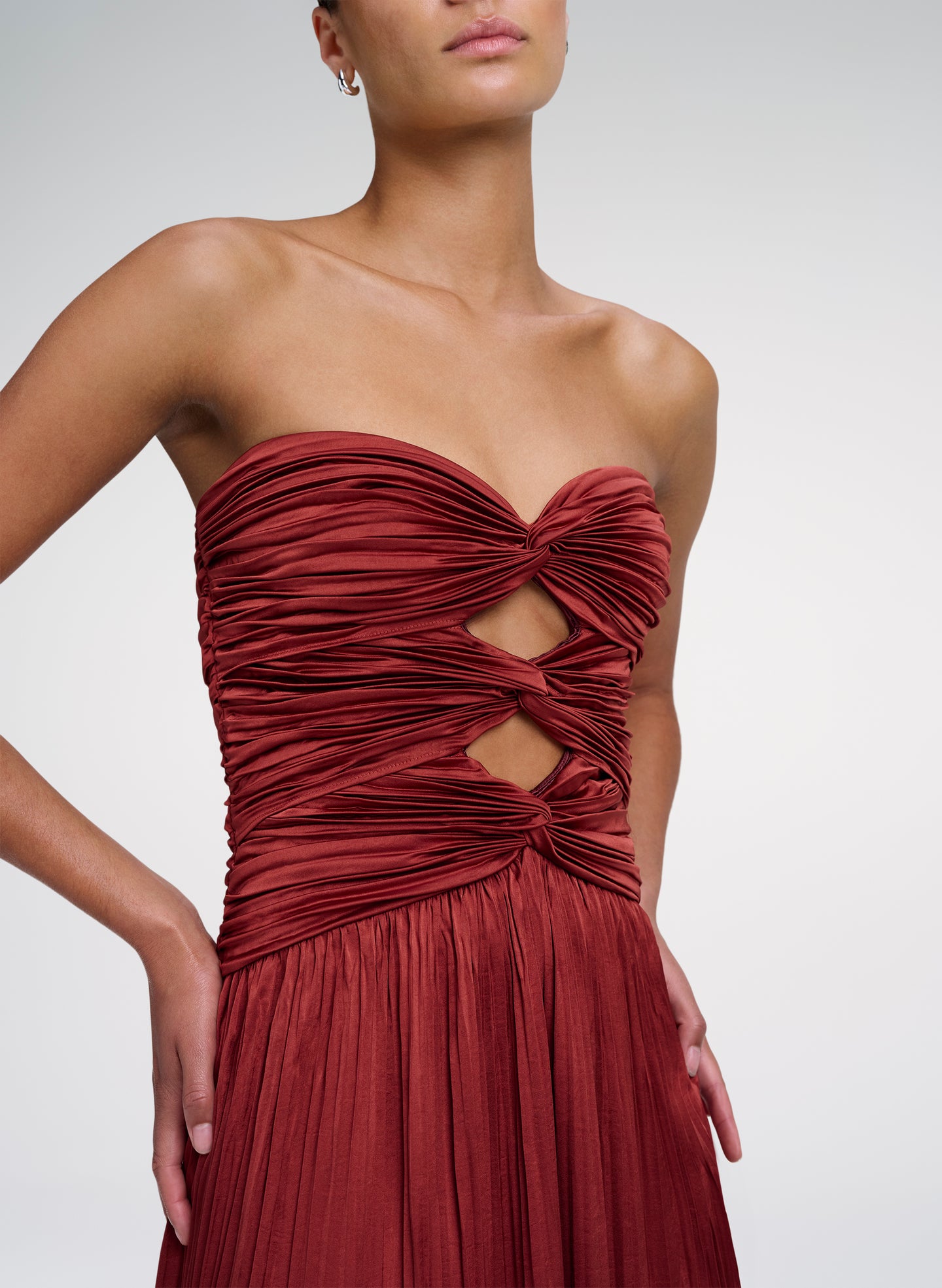 A close-up of a model wearing a strapless deep red dress with a ruched, twisted bodice and a pleated skirt against a light gray studio background.