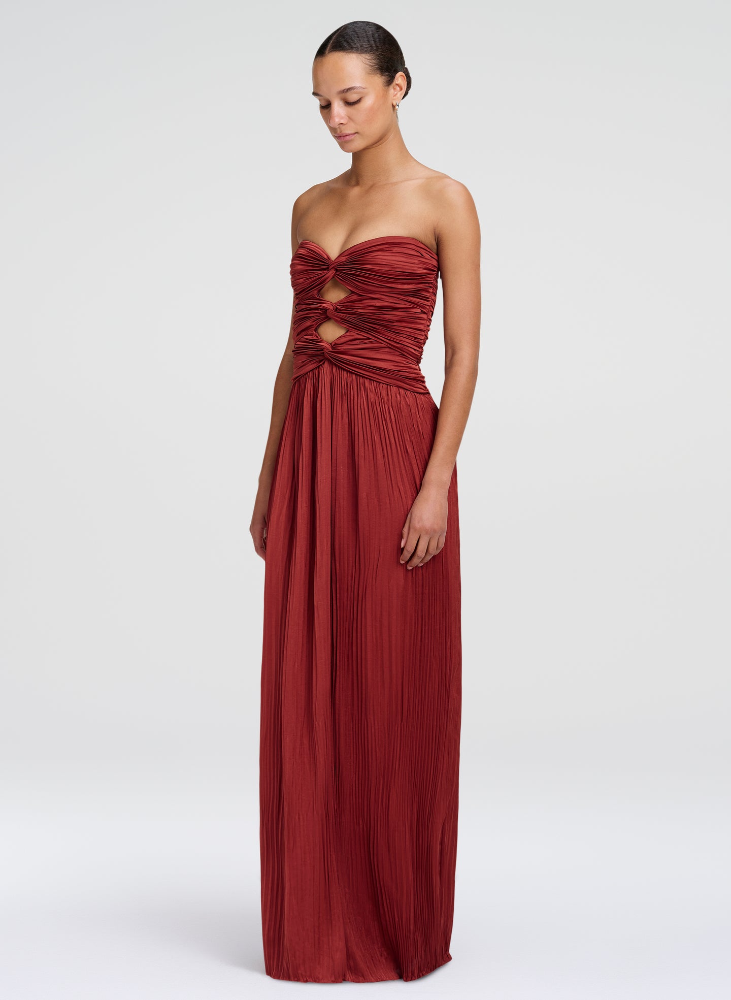A woman modeling a strapless, floor-length burgundy gown with a ruched, twisted bodice and a pleated skirt, photographed against a light gray studio background.