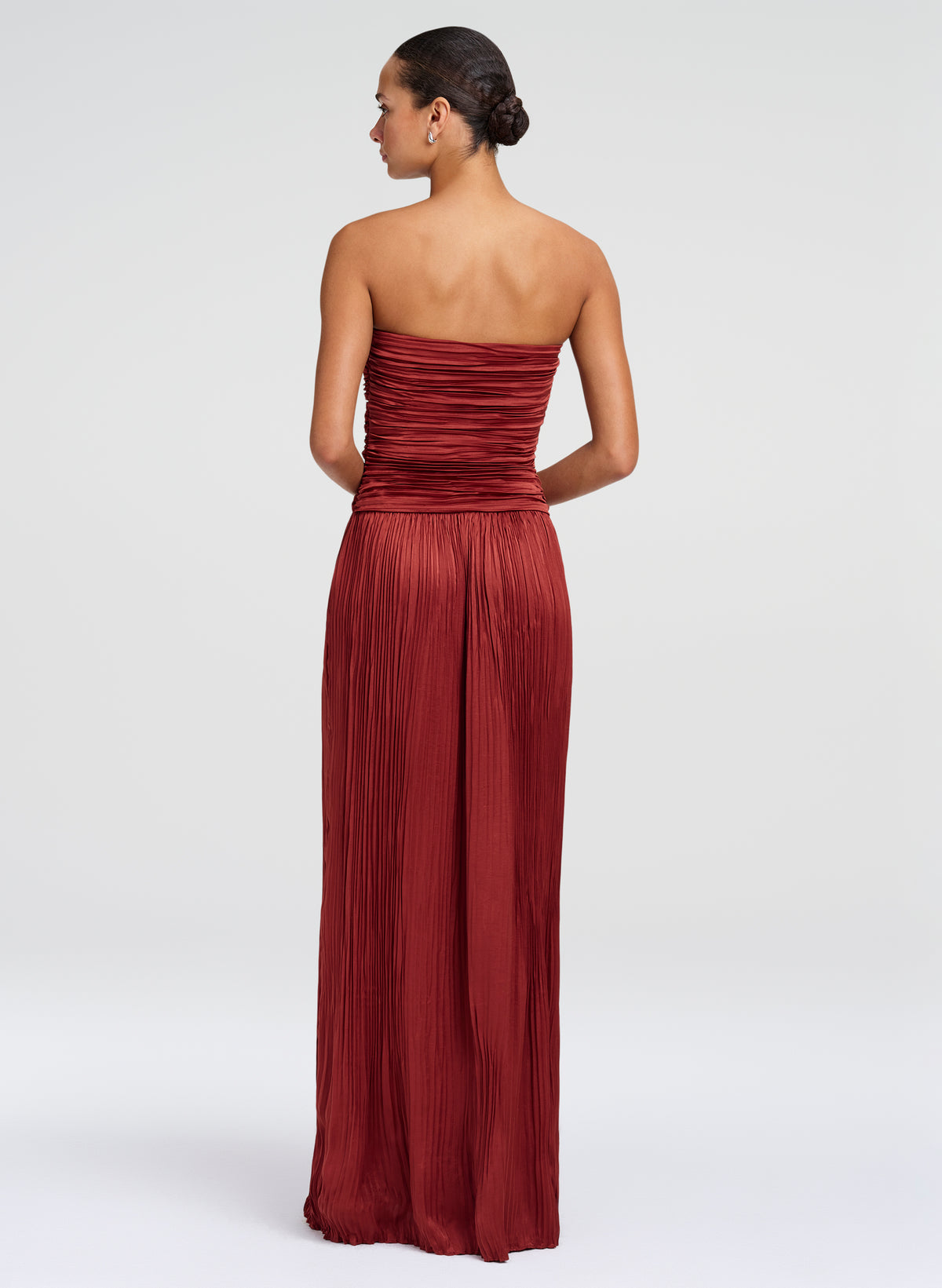 A woman modeling a strapless, floor-length pleated gown from the back, shown against a plain light gray studio background.