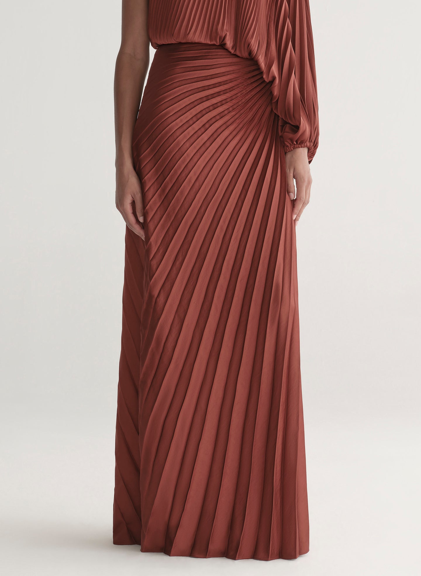 Woman wearing a one-shoulder rust-colored top with matching skirt with pleated details on a white background