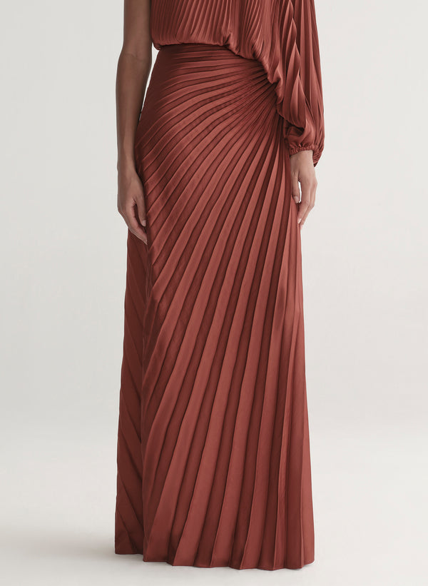 Woman wearing a one-shoulder rust-colored top with matching skirt with pleated details on a white background